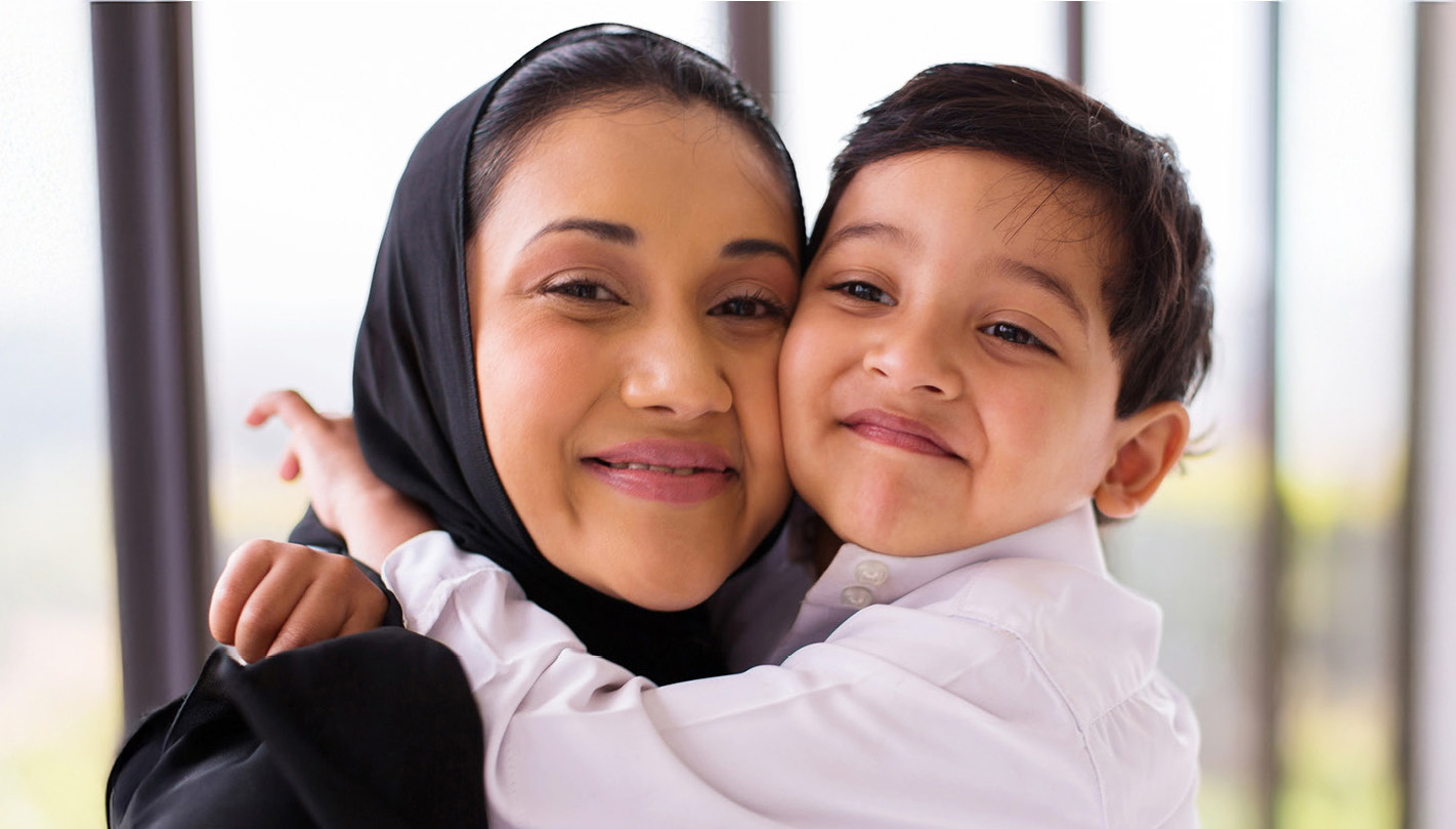 A smiling woman wearing a black headscarf hugs a young child dressed in a white shirt. They are cheek to cheek, both smiling warmly, with soft natural light and a blurred background behind them.